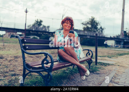 Portrait de femme à la mode senior en utilisant son téléphone portable tout en étant assis sur un banc et d'attendre quelqu'un. Banque D'Images