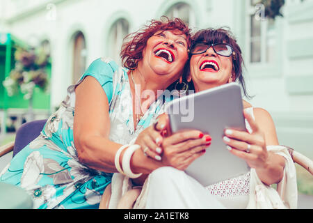 Deux excités senior women sitting in outdoor cafe rire et regarder des médias sociaux et de vidéos drôles sur une tablette. Banque D'Images