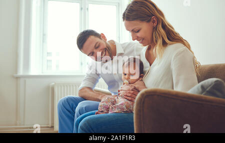 Jeune mère et son père assis sur leur canapé ensemble à la maison, berçant leur adorable petite fille de bébé Banque D'Images