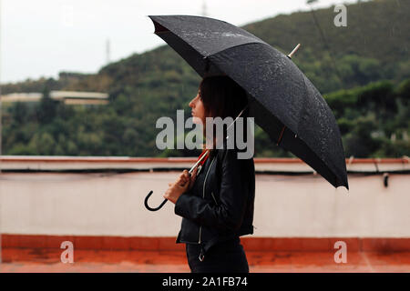 Jeune femme avec un manteau de cuir tient ouverte parapluie noir dans sa main sous la pluie. Le garçon se trouve derrière la montagne. Banque D'Images