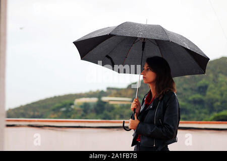 Jeune femme avec un manteau de cuir tient ouverte parapluie noir dans sa main sous la pluie. Le garçon se trouve derrière la montagne. Banque D'Images