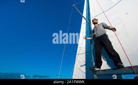 Les jeunes qualifiés positive marin mâle avec barbe portant des lunettes de réglage de la voile sur le bateau et l'examiner tout en fixant les voiles sur flèche Banque D'Images