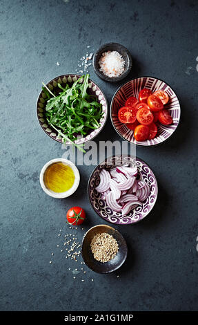 Des ingrédients frais pour la salade, tomates cerises juteuses, de roquette, l'oignon rouge, l'huile de lin et les graines de sésame dans de petits bols de céramique sur pierre sombre backgroun Banque D'Images