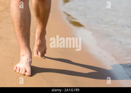 Homme avec des jambes poilues va pieds nus sur la plage de sable le long de la côte près de l'eau avec des vagues Banque D'Images