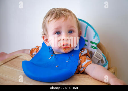 A 1 ans Caucase blond bébé garçon avec un visage fait un gâchis alimenté dans sa chaise haute avec un plastique bleu pelican bavette comme il apprend à manger Banque D'Images