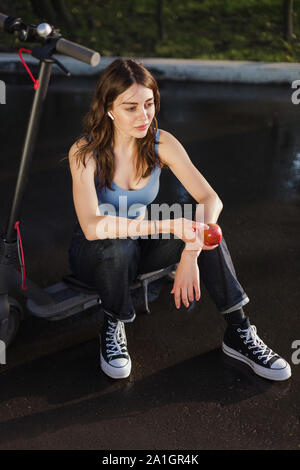 Une fille brune est assise sur un scooter électrique dans le parc avec une pomme dans sa main Banque D'Images