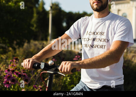 Un homme en blanc T-shirt tient le bras de son scooter électrique tout en équitation dans park Banque D'Images