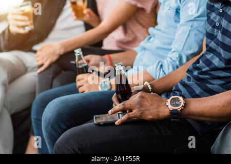 Cropped shot of young African man wearing montre-bracelet élégant, tenant dans la main avec une bouteille de boisson gazeuse et de l'utilisation de téléphone mobile, assis sur le canapé avec fri Banque D'Images