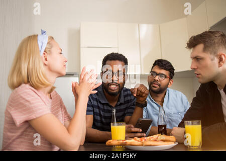 Divers Groupe d'amis assis à table, de manger de la pizza, boire du jus, à l'aide de téléphone mobile, friendly de parler. Les étudiants multiculturels déjeunant à Banque D'Images