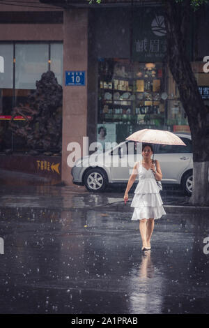 Chengdu, Chine - Juillet 2019 : Belle femme chinoise dans une suite nuptiale robe blanche marcher avec un parapluie lors de la pluie de mousson tropicale Banque D'Images