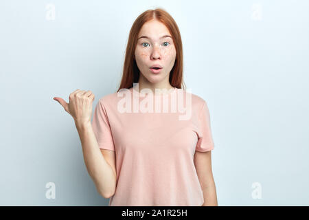 Choqué peur belle jeune femme avec des taches de rousseur foxy longue ligne droite indique avec le pouce de l'espace, acopy montre quelque chose d'étrange à part, isolé sur whi Banque D'Images