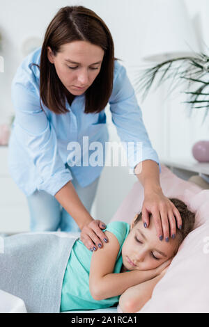 Jeune mère attentive dans casualwear penché au-dessus de sa petite fille malade au lit tandis que les deux en attente de médecin Banque D'Images