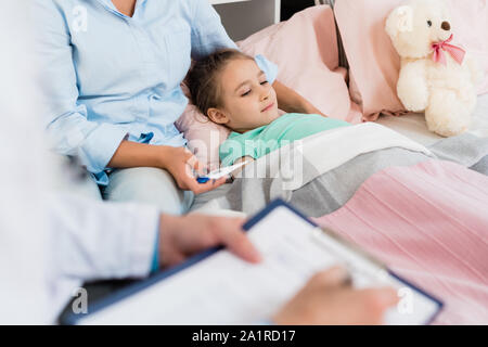 Petite fille malade au lit sous une couverture pendant que sa mère holding thermomètre et médecin décisions ordonnances Banque D'Images