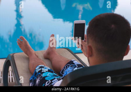 Jeune beau gars s'allonge sur une chaise longue près de la piscine et est titulaire d'un smartphone dans ses mains. Par l'eau. selfies Pour la mémoire de la photo Banque D'Images