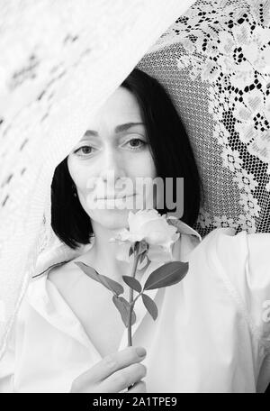 Portrait d'une jeune femme avec une rose blanche dans ses mains, entouré de rideaux blancs en dentelle, noir et blanc tinting Banque D'Images