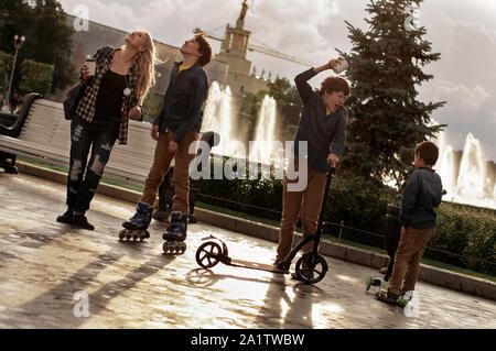 Les frères jumeaux avec un petit frère et maman ride un scooter et de patins dans le parc. Mode de vie sain. Famille Sport Banque D'Images