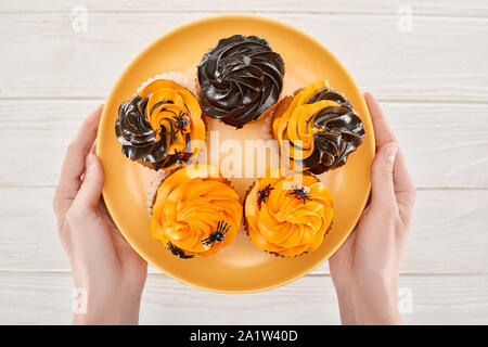 Vue partielle de woman holding delicious cupcakes avec araignées sur la plaque orange sur table en bois blanc, à l'Halloween Banque D'Images