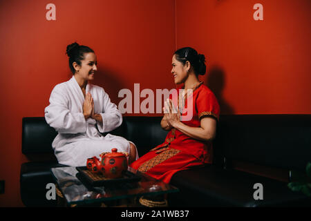Femme souriante et masseur asiatique assis sur un canapé et montrant les mains de souhaits Banque D'Images