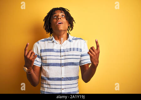 Avec des dreadlocks homme portant chemise rayée sur fond jaune isolé étonné et surpris de la recherche de pointe et avec les doigts et Banque D'Images