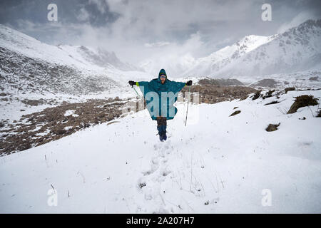 Randonneur en poncho imperméable bleu saute dans les montagnes enneigées. Outdoor travel concept Banque D'Images