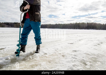 Pêcheur sur glace utilisant un alésage sur le lac en hiver sur une journée ensoleillée dans les bois Banque D'Images