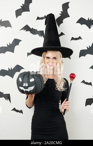 Jeune belle Slim noir sorcière posant avec Halloween citrouille et baguette magique contre mur avec des chauves-souris noir Banque D'Images