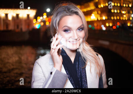 Jeune femme élégante dans un élégant manteau beige utilise un smartphone, sur la rue de la ville la nuit. Belle blonde à parler au téléphone. Banque D'Images