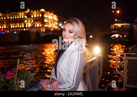 Jeune femme élégante et raffinée dans un manteau beige sur le front de mer de la ville. À l'automne de la ville de nuit. Portrait de charmante blonde de nuit à l'extérieur. Banque D'Images