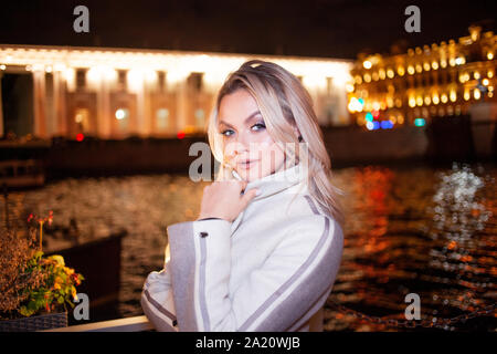 Jeune femme élégante et raffinée dans un manteau beige sur le front de mer de la ville. À l'automne de la ville de nuit. Portrait de charmante blonde de nuit à l'extérieur. Banque D'Images