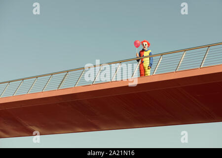 Un clown effrayant colorés portant un jaune, rouge et bleu costume, tenant un ballon rouge dans sa main, debout dans un pont en plein air Banque D'Images