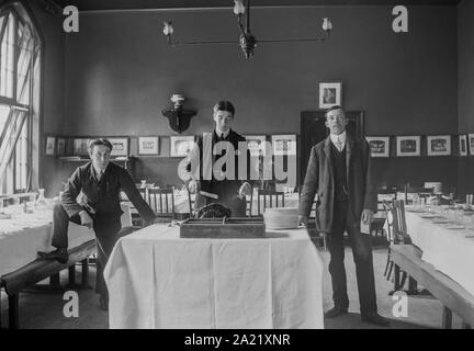 Trois hommes se tenir dans une salle à manger vide, peut-être un club de sport, centre face caméra man carving un joint de la viande. Tables fixées avec de la vaisselle - circa 1900 Banque D'Images