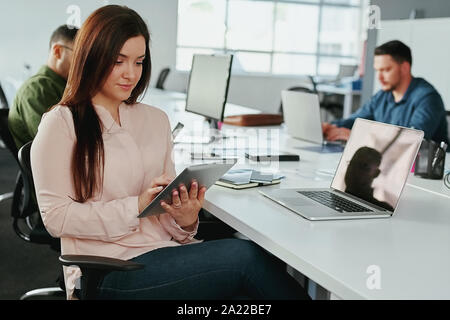Un séduisant jeune smiling businesswoman using digital tablet avec des collègues travaillant au bureau en arrière-plan à l'office de tourisme - Office de démarrage Banque D'Images