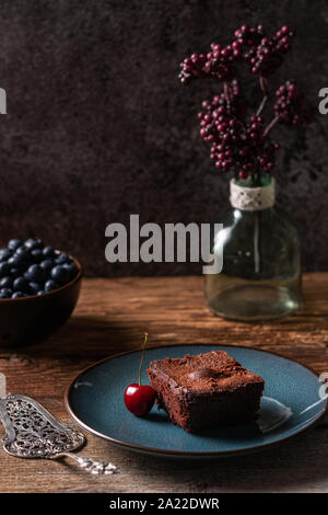 Brownie au chocolat vegan fait maison, et fraîchement cuits servis sur une plaque bleue avec des bleuets frais et d'une cerise. Disposés sur une table en bois avec bac Banque D'Images