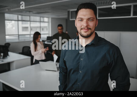 Portrait d'un jeune businessman smiling and looking at camera tandis que d'autres business people talking in office Banque D'Images