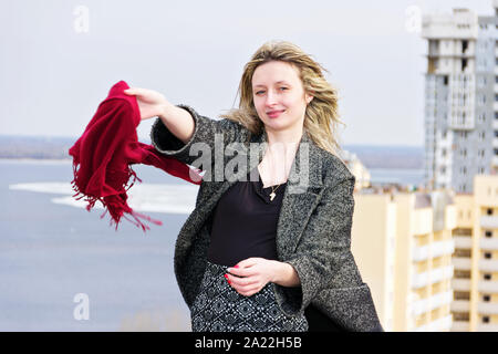 Fille blonde dans un manteau gris tenant un foulard rouge sur le toit d'un gratte-ciel. Banque D'Images