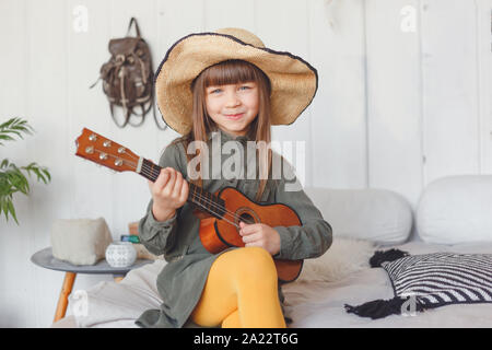 Kid girl in hat jouer ukulele à la maison Banque D'Images