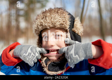 Portrait d'un enfant garçon heureux qui sort sa langue s'amusant dans la neige durant la saison d'hiver et portant un chapeau garni de fourrure Banque D'Images