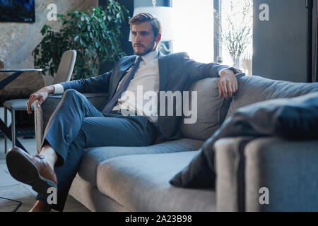 Jeune homme charmant et pense à business tout en étant assis sur le canapé dans son bureau moderne. Banque D'Images