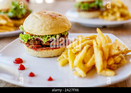 Burger de boeuf avec patty, laitue, bacon, servis avec frites sur une assiette blanche sur une table en bois. Deux autres burgers sur l'arrière-plan. Banque D'Images