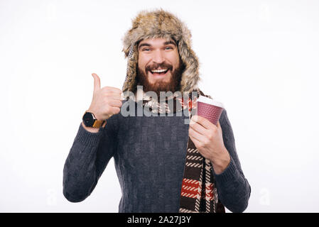 Bon café joyeux hipster barbu portant foulard et bonnet hiver montrant comme geste tout en tenant une tasse à emporter. Banque D'Images