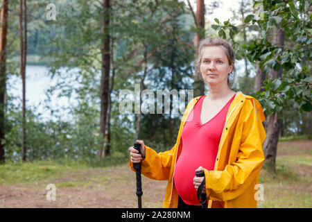 Portrait of young pregnant woman wearing raincoat en été permanent forest holding bâtons appréciant la nature - Grossesse en santé concept de vie Banque D'Images