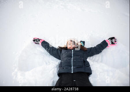 Girl smiling tout en faisant l'ange de la neige dans la neige dans la cour avant Banque D'Images
