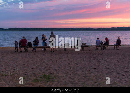 Des gens assis sur des bancs au bord du lac rouge et bleu à regarder le coucher du soleil. Groupe de jeunes gens s'amuser à la plage près du lac. Partie au coucher du soleil. Banque D'Images