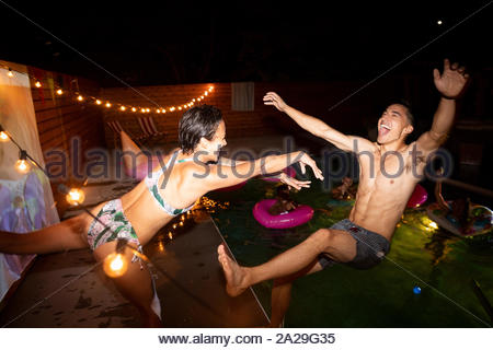 Jeune femme ludique poussant l'homme dans l'été de la piscine de nuit