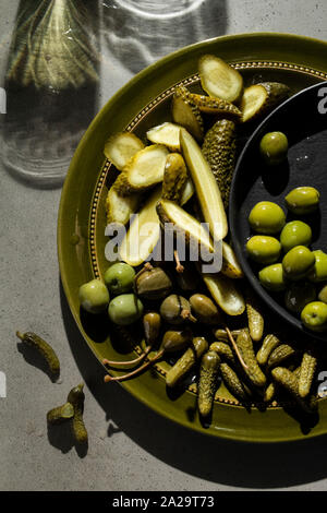 Une sélection de cornichons au vinaigre, olives vertes et câpres petits fruits sur une plaque de vert olive. Ombres sur le fond béton suggèrent verres de vin. Banque D'Images