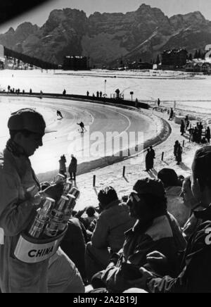 Aux Jeux Olympiques d'hiver de 1956 à Cortina d'Ampezzo les compétitions de patinage de vitesse ont eu lieu sur le lac gelé de Misurina. La photo montre un vendeur de boissons qui purge certains spectateurs dans les tribunes. Dans l'arrière-plan, un 500 mètres est détenu. Banque D'Images