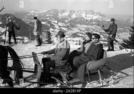 Couple sur une promenade en traîneau, certains skieurs dans l'arrière-plan et une vue sur les Alpes. La photo a été prise à l'occasion de la course du Hahnenkamm en 1962. La course du Hahnenkamm a eu lieu sur le Hahnenkamm de Kitzbühel depuis 1931. Banque D'Images