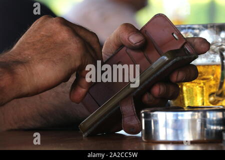 Close up of working man's hands using smart phone in fine en cuir avec partiellement ivre verre de bière et en acier inoxydable cendrier sur le bar en bois haut Banque D'Images