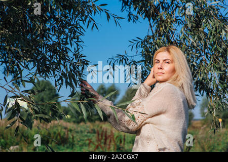 Heureux belle blonde femme vêtue de sweater debout à côté de l'arbre avec des feuilles vertes brunchs holding. Banque D'Images