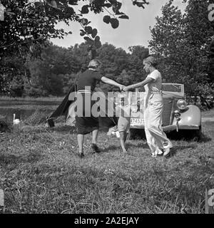 Zwei junge Frauen tanzen Ringelreihen mit einem kleinen Jungen vor einem Ford V8 733Am Ufer eines voit, Deutschland 1930er Jahre. Deux jeunes femmes danser ring-a-ring-a-roses wit ha petit garçon en face d'une Ford V8 décapotable, Allemagne 1930. Banque D'Images